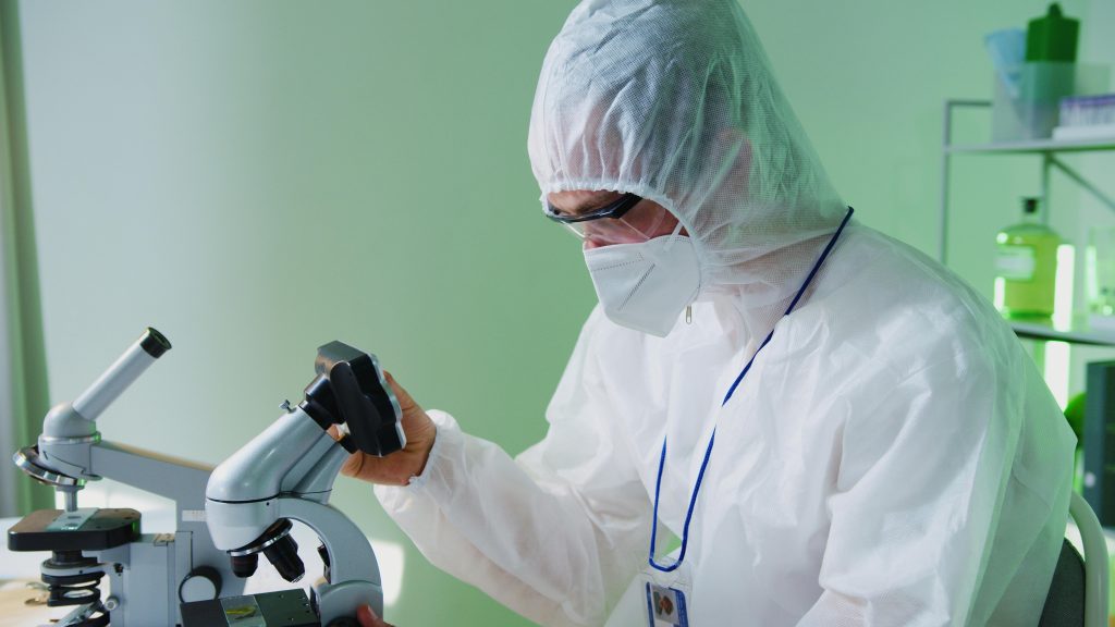 A scientist in protective gear examines samples under a microscope in a lab.